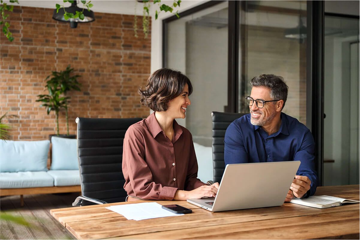 Two smiling colleagues, a woman in a maroon shirt and a man in a navy blue shirt, sit at a wooden table in a modern office space, working together on a laptop.
